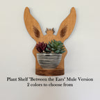 Wooden mule-shaped plant shelf with long ears and a striped ceramic pot holding succulents, shown against a plain light background with text reading 'Plant Shelf "Between the Ears" Mule Version 2 colors to choose from'.
